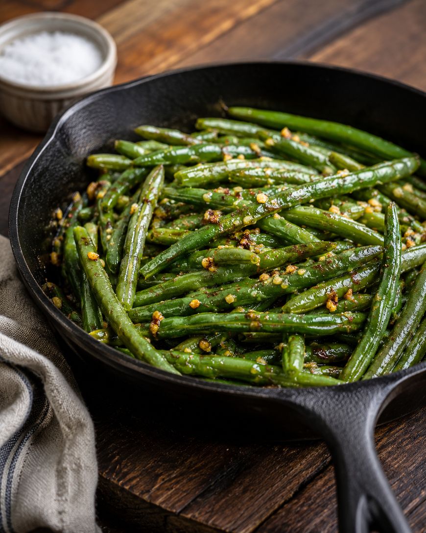 A cast iron skillet filled with sautéed green beans topped with minced garlic sits on a wooden table. A bowl of salt and a beige napkin are nearby.