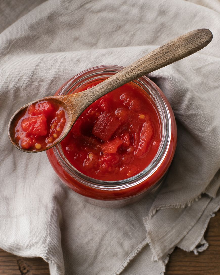 An open glass jar of chunky tomato sauce sits on a beige cloth, with a wooden spoon resting on top holding a serving of the sauce.