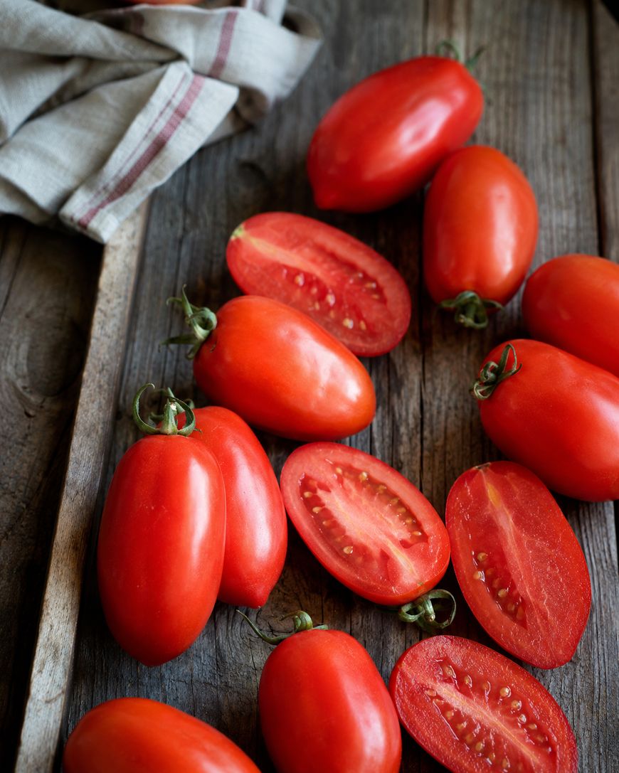 Several whole and sliced Roma tomatoes are arranged on a rustic wooden surface next to a beige cloth with red stripes.