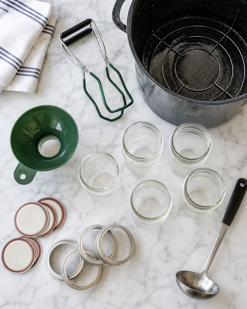 Canning supplies on a marble countertop, including empty glass jars, lids, metal rings, a ladle, jar lifter, green funnel, large pot with rack, and a striped dish towel.