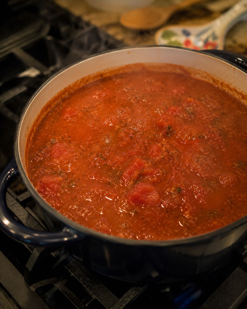 A large dark blue pot sits on a stovetop, filled with simmering red tomato sauce with visible herbs and tomato chunks. A spoon and dishes are in the background.
