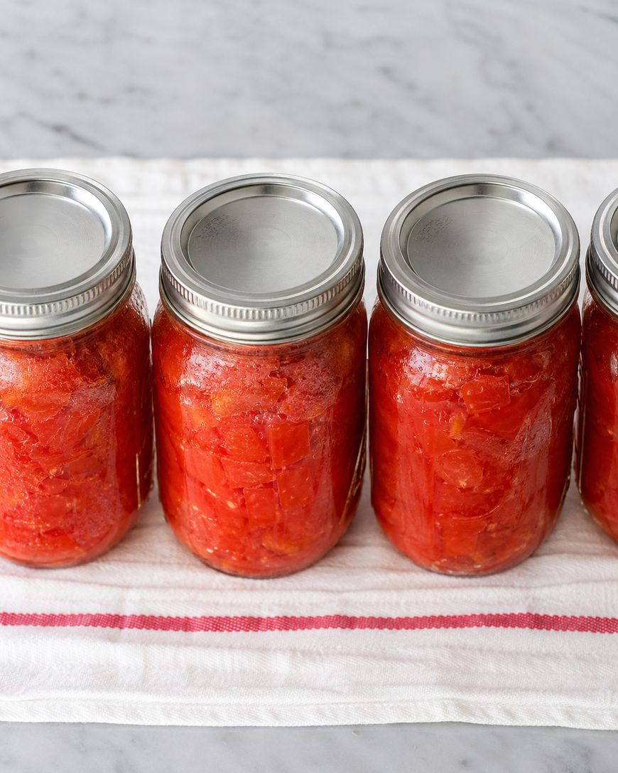 Four glass jars filled with diced tomatoes are lined up on a white cloth with a red stripe, set on a marble countertop. The jars are sealed with metal lids.