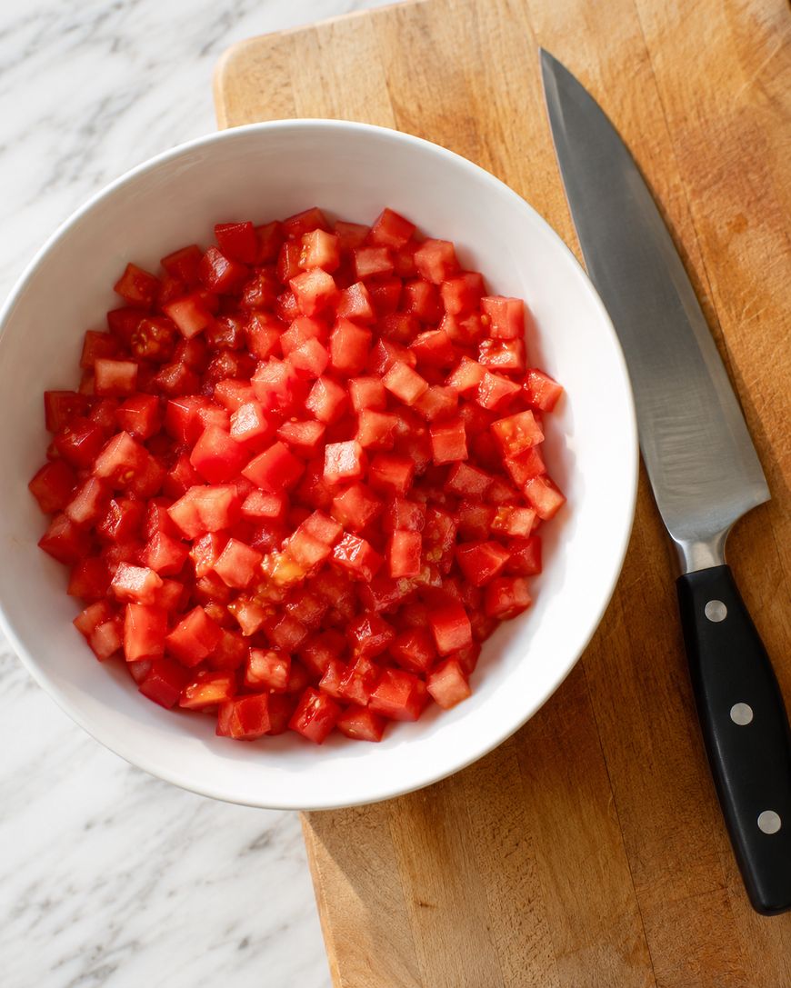 A white bowl filled with diced tomatoes sits on a wooden cutting board next to a chef’s knife with a black handle.