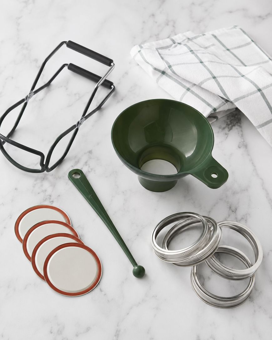 Canning tools on a marble surface: a green funnel, jar lifter tongs, a checkered towel, a bubble remover stick, metal lids, and metal rings for canning jars.