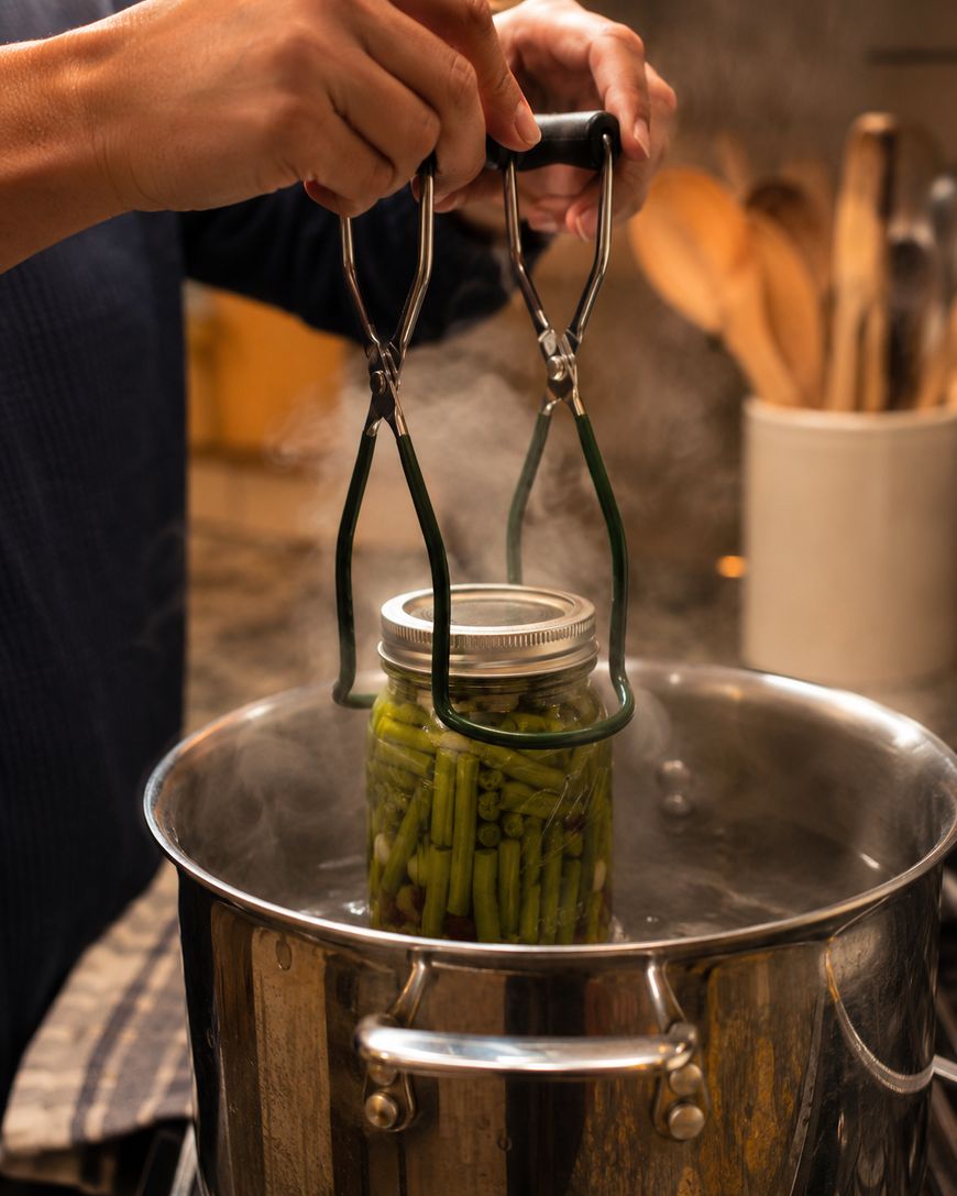 A person uses jar tongs to place a glass jar filled with green beans into a steaming pot of boiling water, likely for the process of home canning or preserving vegetables in a kitchen setting.