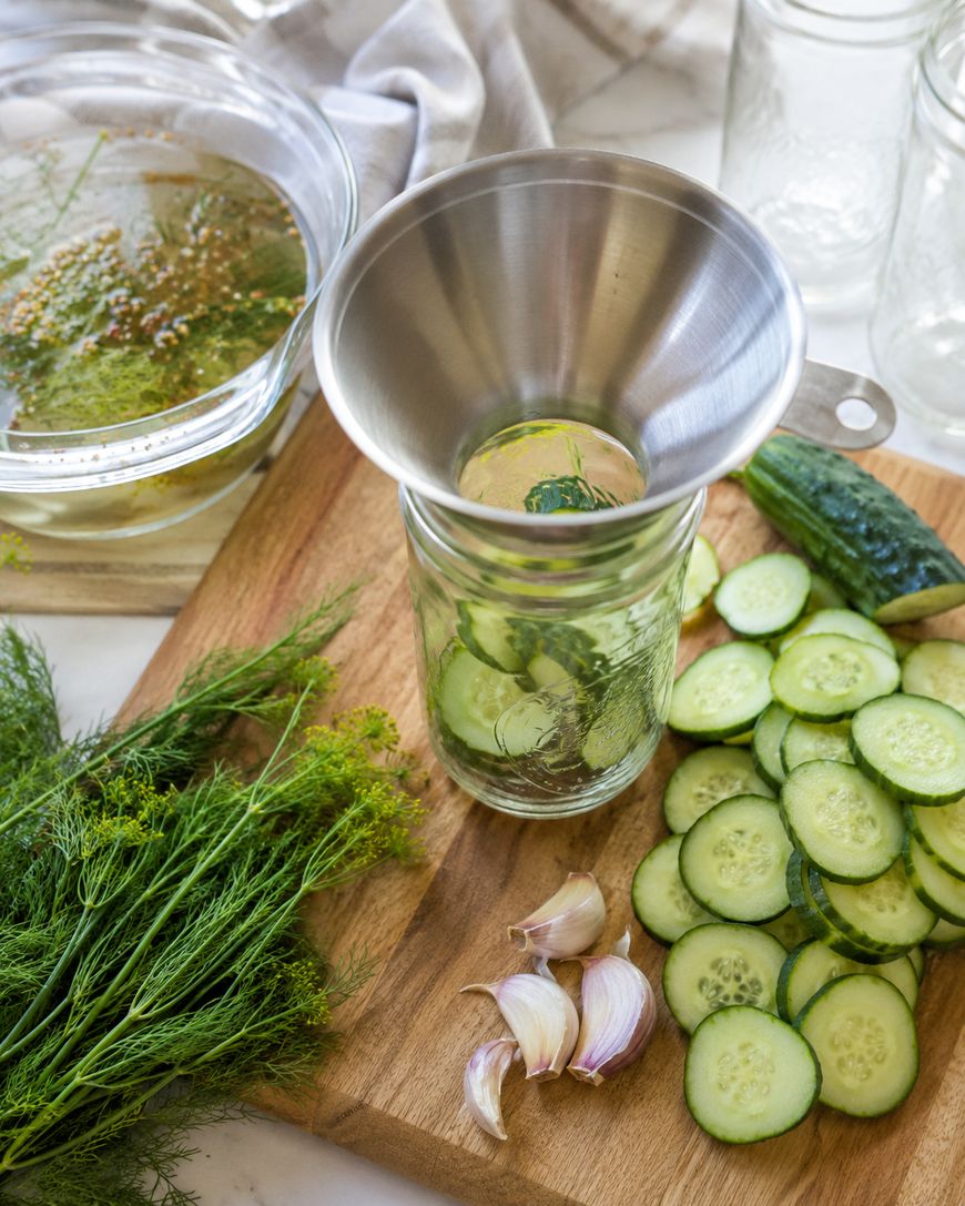 Sliced cucumbers, garlic cloves, and fresh dill on a wooden cutting board next to a glass jar with a metal funnel, ready for pickling. A bowl of pickling brine and empty jars are in the background.