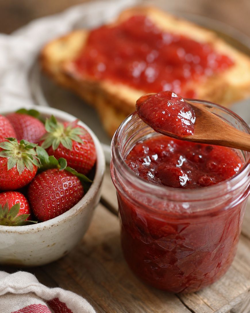A jar of strawberry jam with a wooden spoon, a bowl of fresh strawberries, and a slice of toast spread with jam on a plate in the background, all on a rustic wooden table.