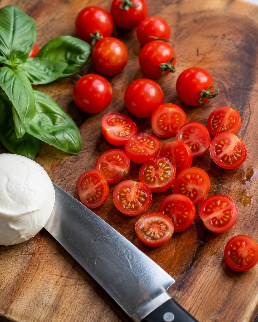 Fresh basil leaves, whole and halved cherry tomatoes, a ball of mozzarella cheese, and a chef’s knife are arranged on a wooden cutting board.