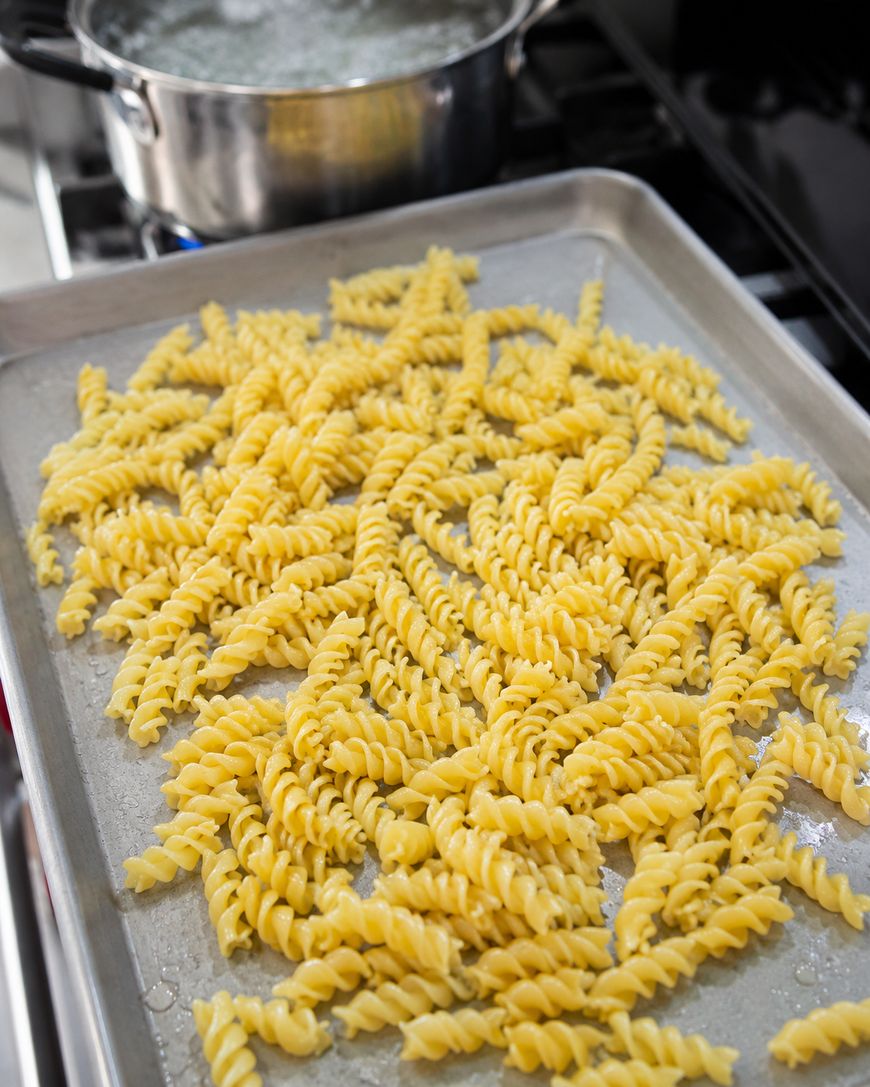 A baking sheet covered with cooked fusilli pasta sits in the foreground, while a pot of boiling water is visible on the stove in the background.