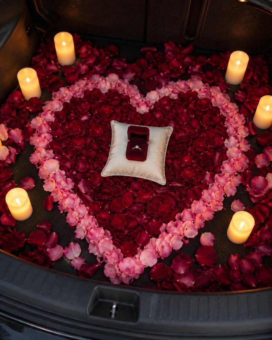 A heart made of red and pink rose petals is arranged in the trunk of a car, surrounded by six lit candles. A ring box sits on a small pillow at the center of the heart.