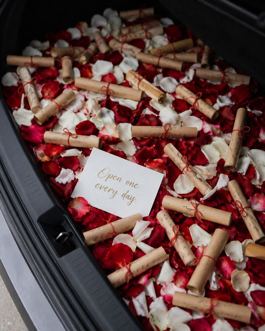 An open car trunk filled with red and white rose petals and rolled-up notes tied with red string. In the center, a card reads, Open one every day.