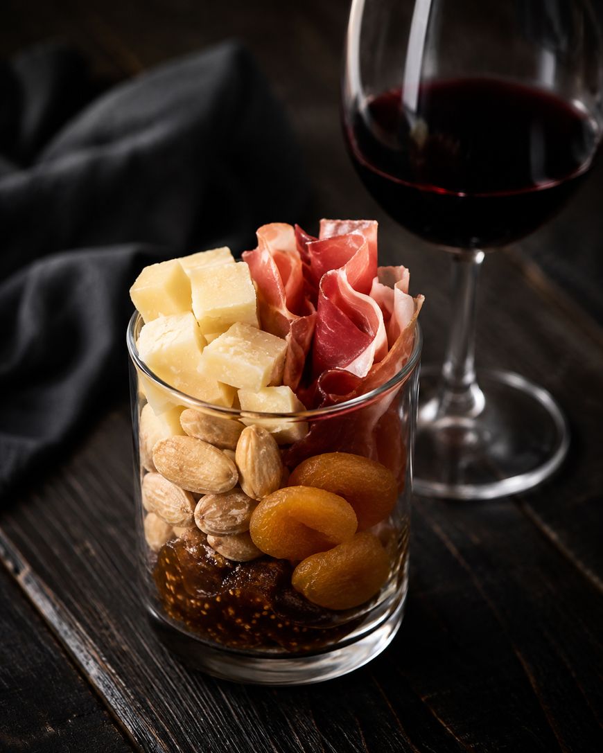 A glass filled with cubed cheese, prosciutto, almonds, dried apricots, and fig spread sits on a dark wooden table next to a glass of red wine, with a black napkin in the background.