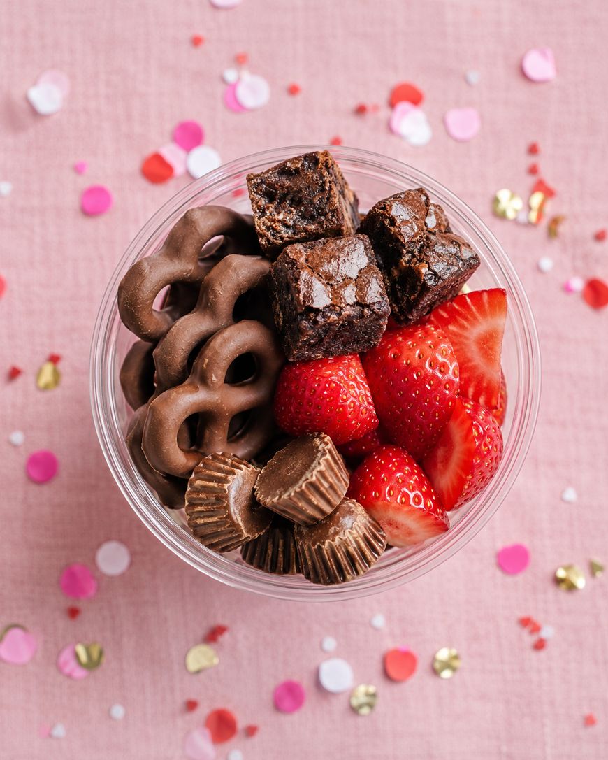 A clear cup filled with chocolate-covered pretzels, mini peanut butter cups, brownie bites, and fresh strawberries sits on a pink surface decorated with colorful confetti.
