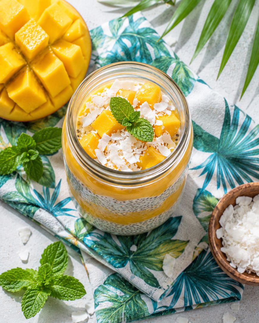 A glass jar filled with chia pudding, topped with mango cubes, coconut flakes, and a mint sprig, sits on a tropical-patterned cloth. Sliced mango, coconut flakes, and mint leaves are nearby.