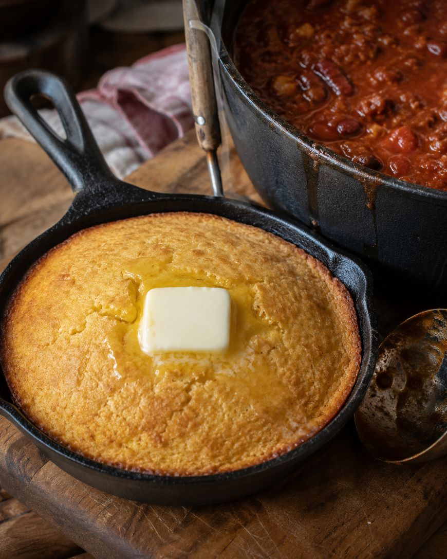 A skillet of golden cornbread with a pat of melting butter on top sits on a wooden board next to a pot of chili. A spoon rests nearby, and a cloth is partially visible in the background.