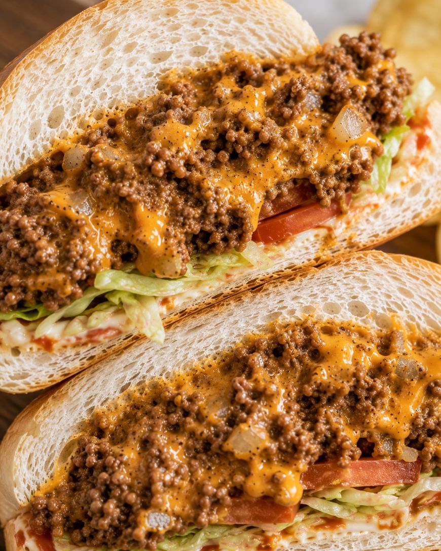 Close-up of two sandwiches filled with shredded lettuce, sliced tomatoes, and topped with seasoned ground beef and melted orange cheese sauce on white sub rolls.