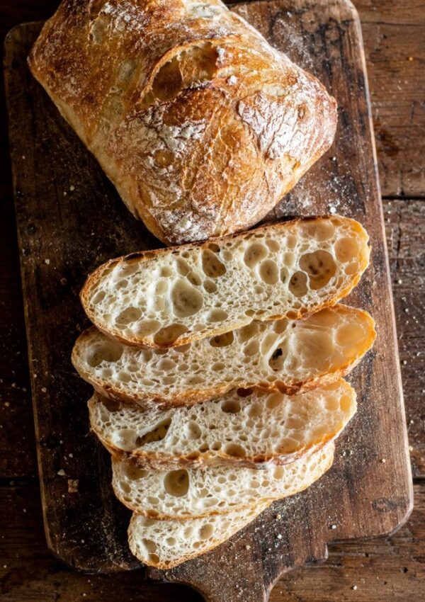 A loaf of rustic ciabatta bread, partially sliced, sits on a wooden cutting board. This artisan bread boasts a golden, crusty exterior and airy, hole-filled slices—just like the best ciabatta bread recipe would yield.