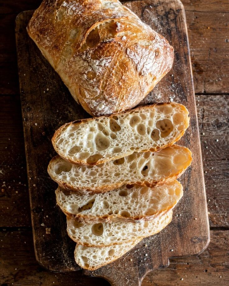 A loaf of rustic ciabatta bread, partially sliced, sits on a wooden cutting board. This artisan bread boasts a golden, crusty exterior and airy, hole-filled slices—just like the best ciabatta bread recipe would yield.