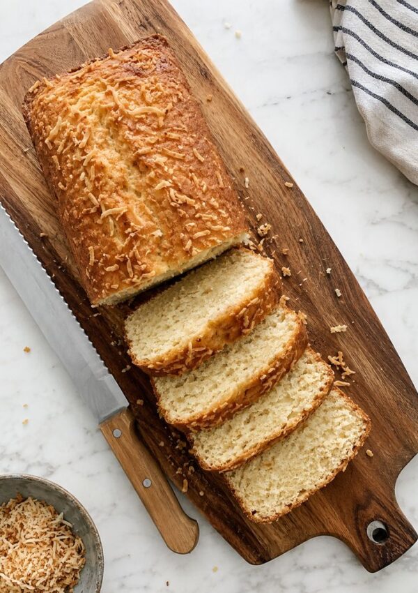A loaf of coconut bread recipe topped with shredded cheese is sliced on a wooden cutting board next to a bread knife. A striped towel and a small bowl of toasted shredded cheese are also on the marble surface nearby.