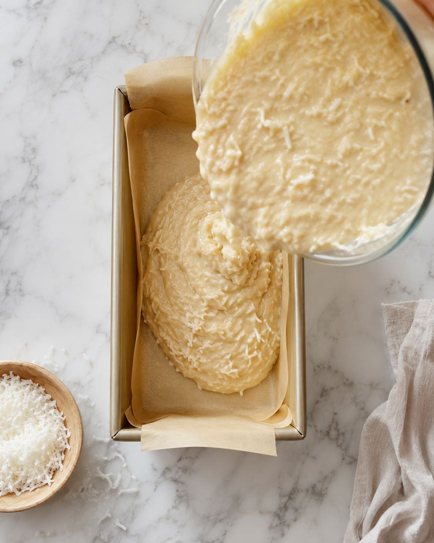 A glass bowl pours cake batter with shredded coconut into a parchment-lined loaf pan. Nearby, a small wooden bowl with shredded coconut sits on a marble surface, next to a beige cloth.