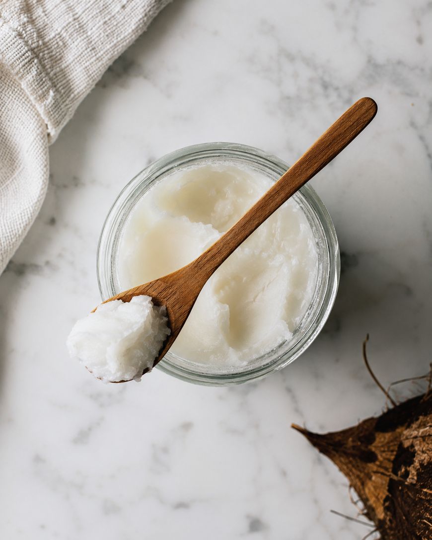 A glass jar filled with solid coconut oil sits on a marble surface. A wooden spoon with a scoop of coconut oil rests on top, and part of a coconut shell is visible in the corner.