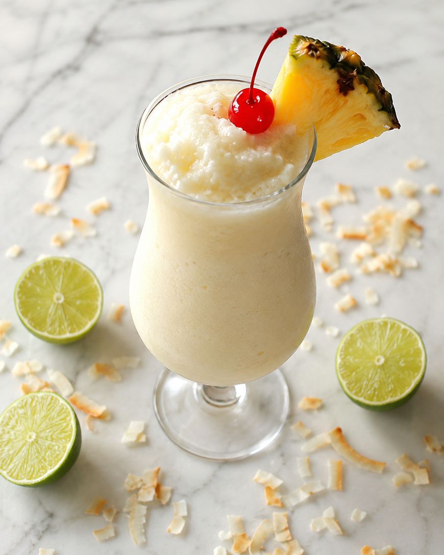 A creamy piƱa colada in a curved glass, garnished with a pineapple wedge and a maraschino cherry, sits on a marble surface surrounded by lime halves and scattered toasted coconut flakes.