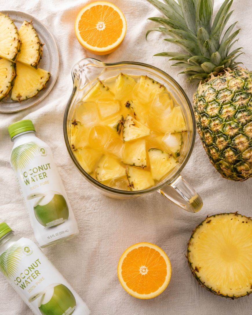 A glass pitcher filled with ice, pineapple chunks, and yellow juice sits on a table, surrounded by coconut water bottles, pineapple slices, a halved orange, and a whole pineapple.