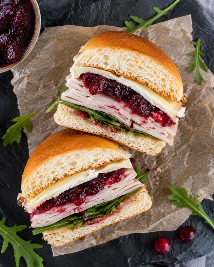 A sandwich cut in half on parchment paper, filled with sliced turkey, cheese, cranberry sauce, arugula, and mayonnaise, with extra arugula and a bowl of cranberry sauce nearby.
