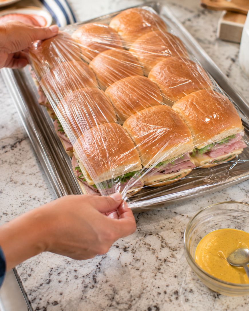 A person wraps a tray of ham and cheese sliders with plastic wrap on a marble countertop; a bowl of yellow mustard sits nearby.