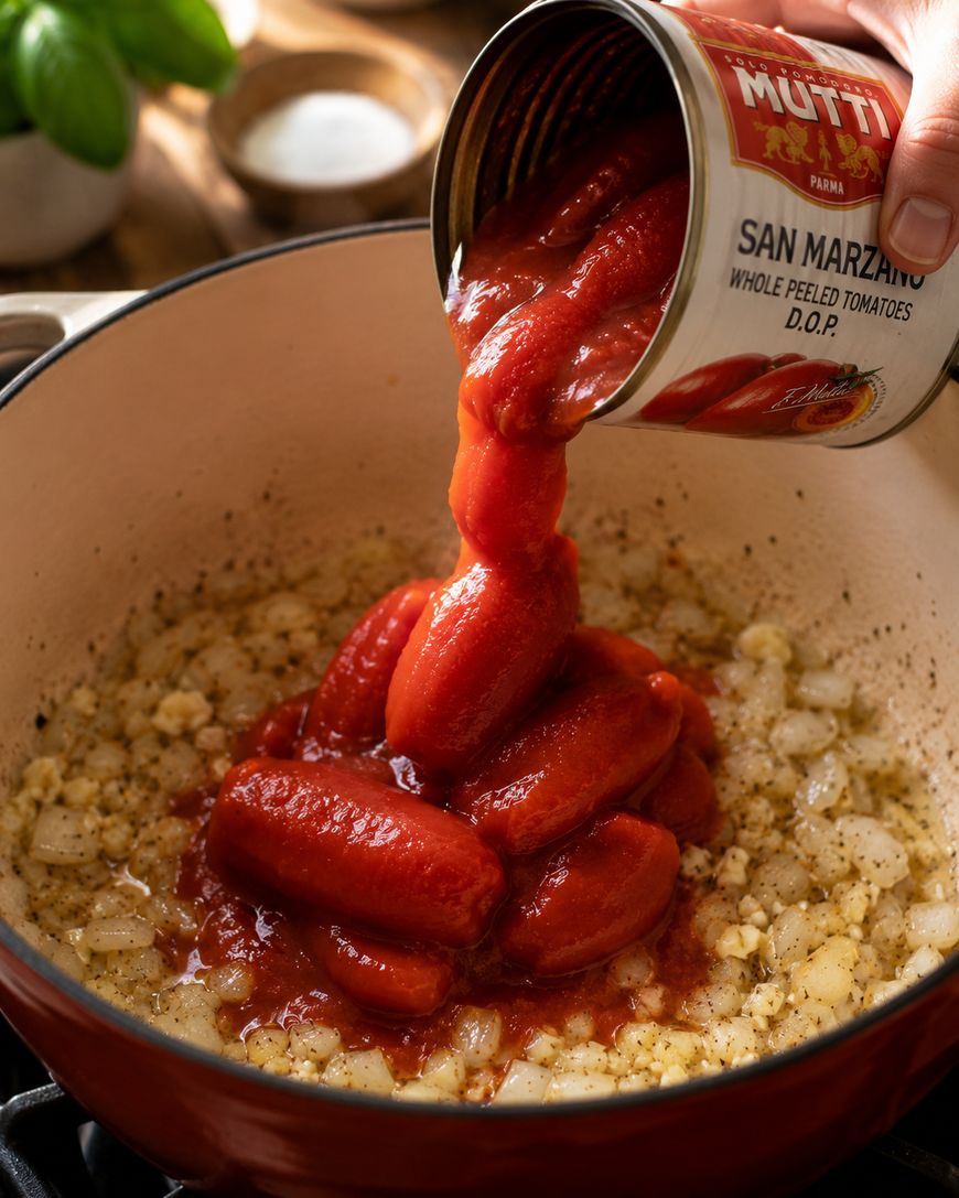 A can of whole peeled San Marzano tomatoes is being poured into a pot with sautéed onions and garlic, ready for cooking.