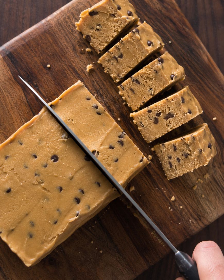 A hand slices a block of chocolate chip cookie dough into even pieces on a wooden cutting board.