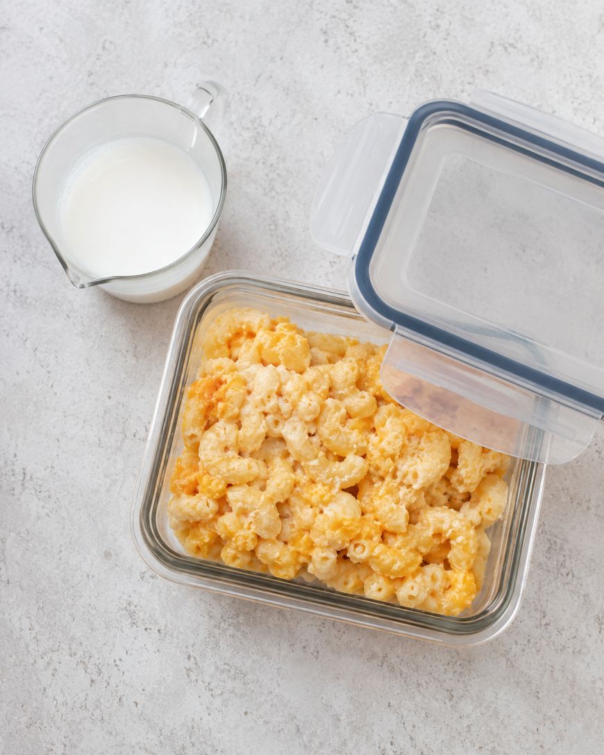 A glass container of baked macaroni and cheese with a partially open lid sits beside a glass measuring cup filled with milk on a light, textured surface.