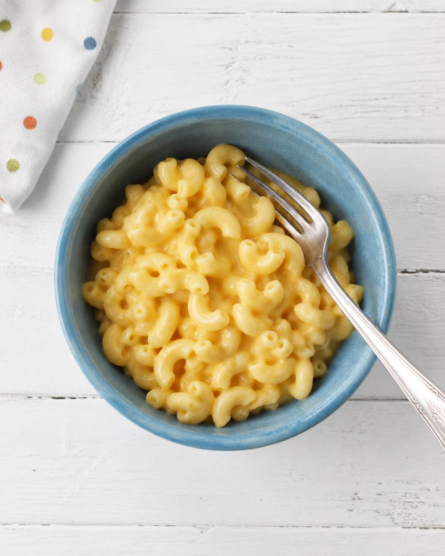 A blue bowl filled with creamy macaroni and cheese sits on a white wooden surface. A silver fork rests on the side of the bowl, and a white napkin with colorful polka dots is partially visible in the corner.