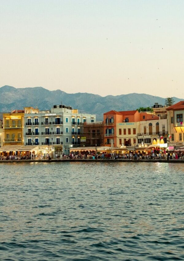 Colorful buildings line a waterfront promenade at sunset, with mountains in the background and calm blue water in the foreground. People gather along the lively shoreline.