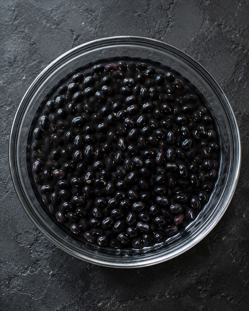 A clear glass bowl filled with black beans sits on a dark textured surface, photographed from above.