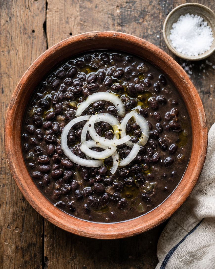A rustic clay bowl filled with black bean soup, topped with sliced raw onions and a drizzle of oil. A small dish of coarse salt and a beige napkin are beside the bowl on a wooden table.