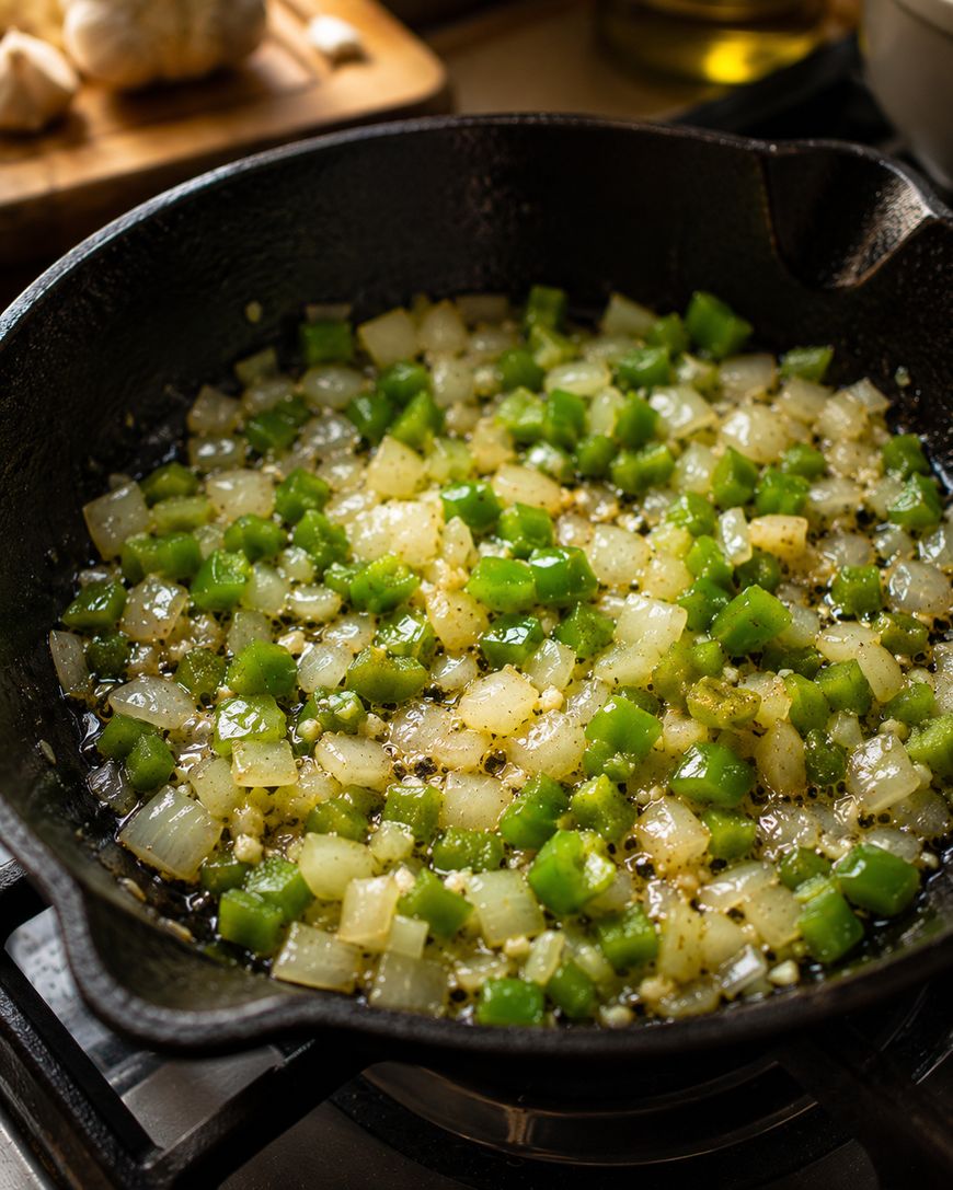 Diced green bell peppers and onions are being sautéed with oil and spices in a black cast iron skillet on a stovetop.