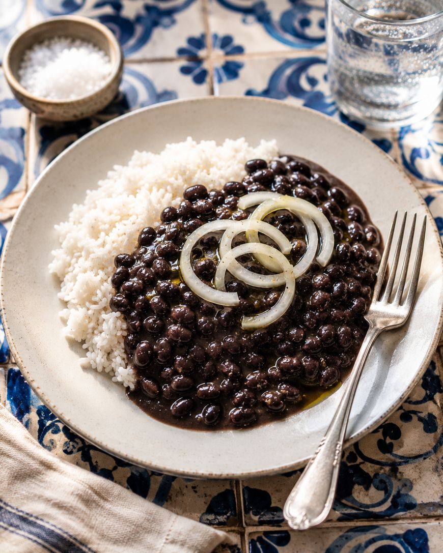 A plate of white rice topped with black beans and garnished with sliced onions, served with a fork on a patterned tile surface. A glass of water and a small bowl of salt are in the background.