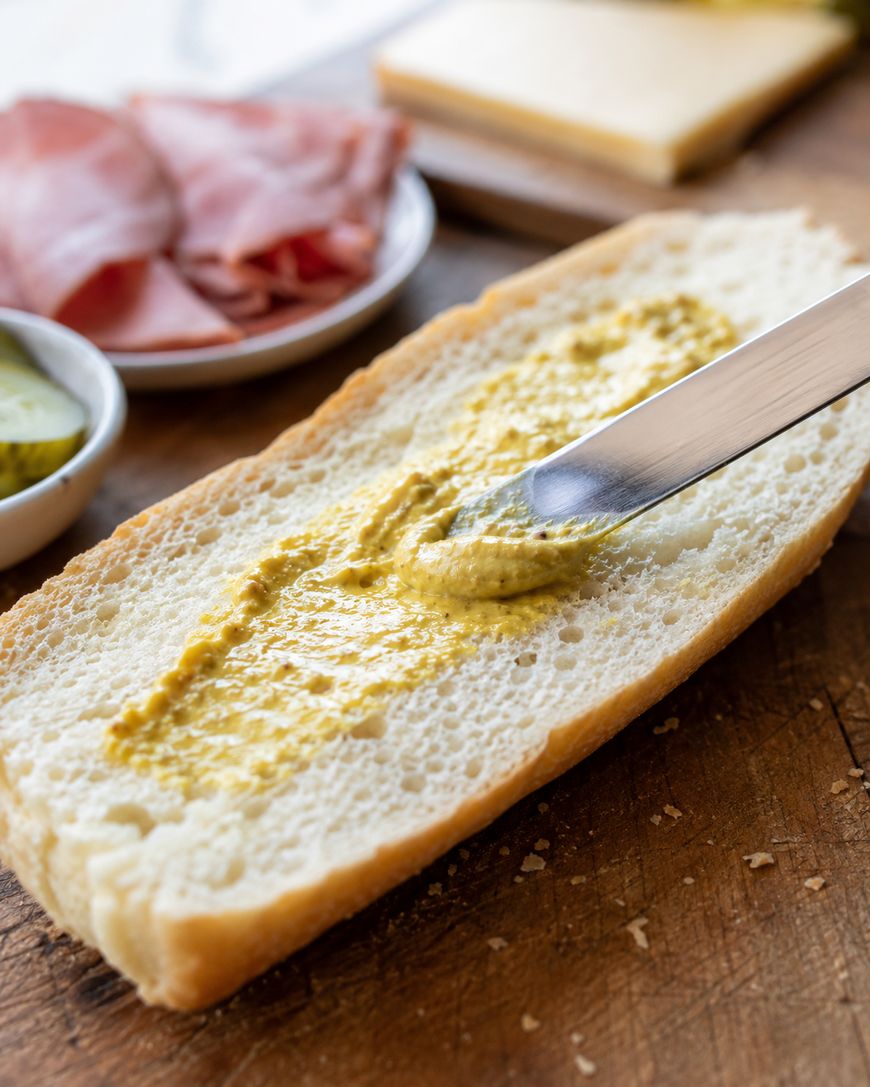 A knife spreads mustard on a slice of fresh bread, with sliced ham, cheese, and pickles in small bowls in the background on a wooden surface.