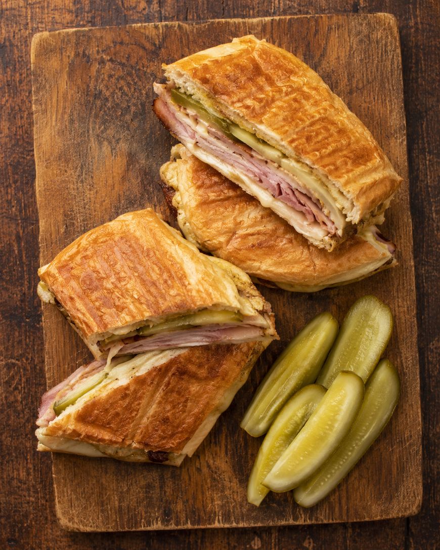 Two halves of a pressed sandwich with ham, cheese, and pickles are on a wooden board, served with several pickle spears on the side.