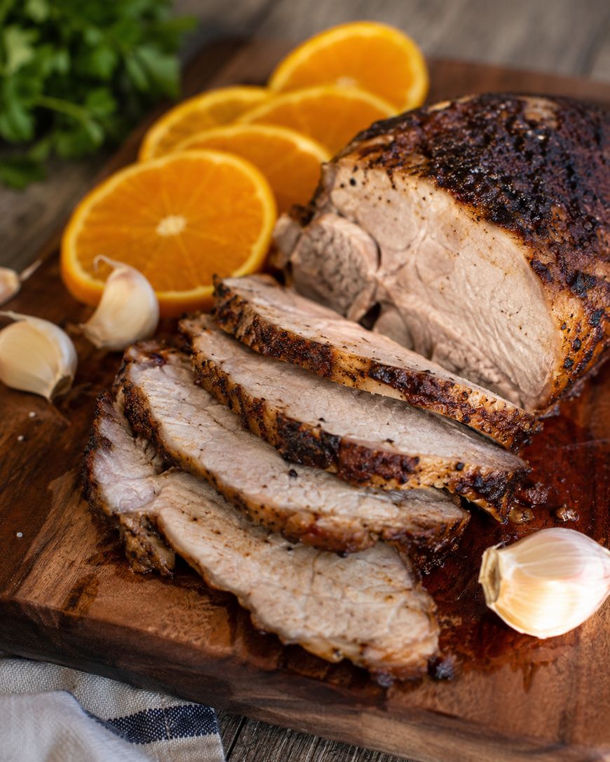 Sliced roasted pork on a wooden cutting board, garnished with whole garlic cloves and orange slices, with fresh parsley in the background.