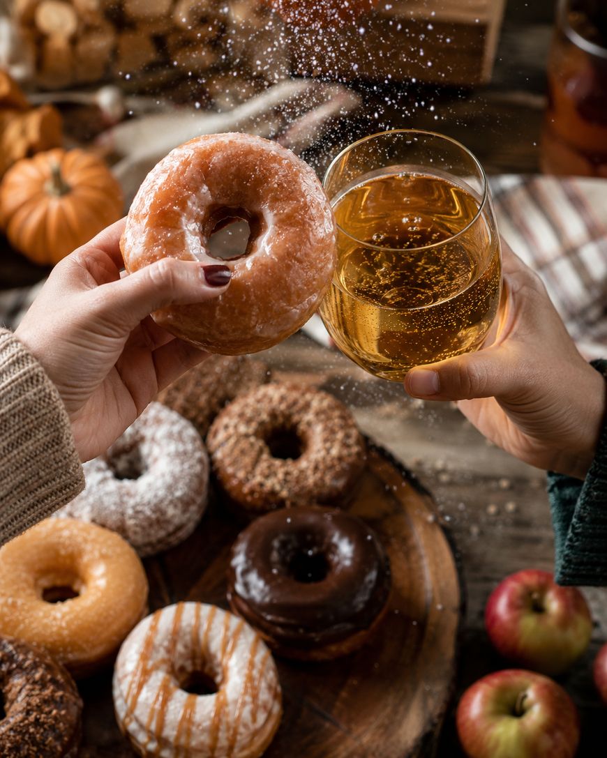 Two hands hold a glazed donut and a glass of apple cider in a toast, with powdered sugar falling. A wooden table below displays assorted donuts and apples, creating a cozy autumn atmosphere.