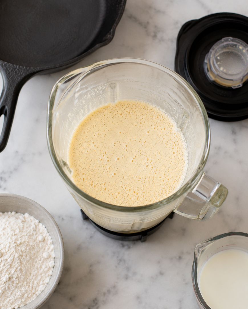 A blender filled with a pale, frothy batter sits on a marble countertop. Nearby are a bowl of flour, a small pitcher of milk, and a cast iron skillet, suggesting ingredients for a recipe.