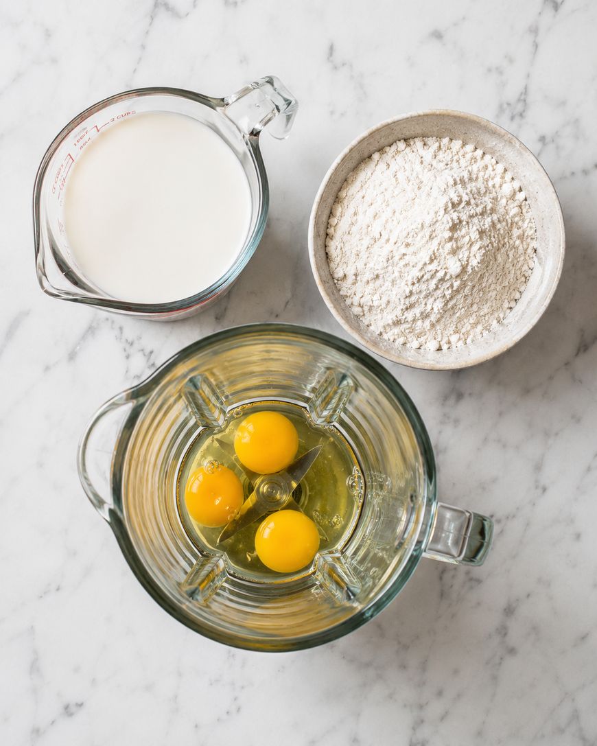 A glass measuring cup with milk, a bowl of flour, and a blender containing three cracked eggs sit on a white marble countertop.