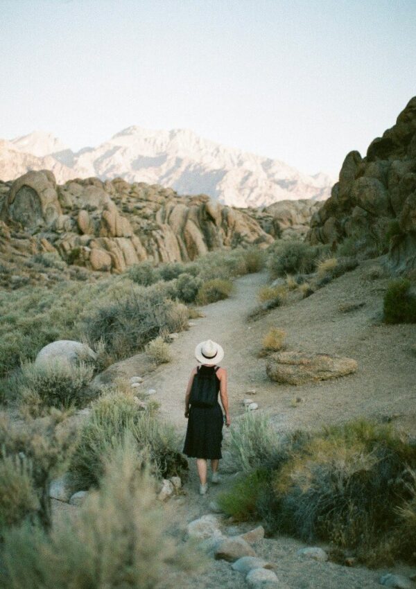 A person in a black dress and straw hat walks alone on a dirt trail through a rocky desert landscape surrounded by shrubs, with distant mountains visible under a clear sky.
