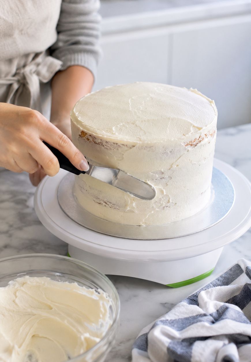 A person uses an offset spatula to spread white icing on a round cake placed on a rotating cake stand, with a bowl of frosting and a striped towel on a marble countertop nearby.