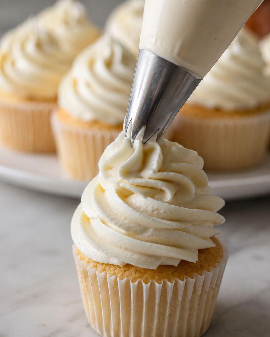 A close-up of a cupcake being topped with swirled vanilla frosting from a piping bag, with more frosted cupcakes blurred in the background.