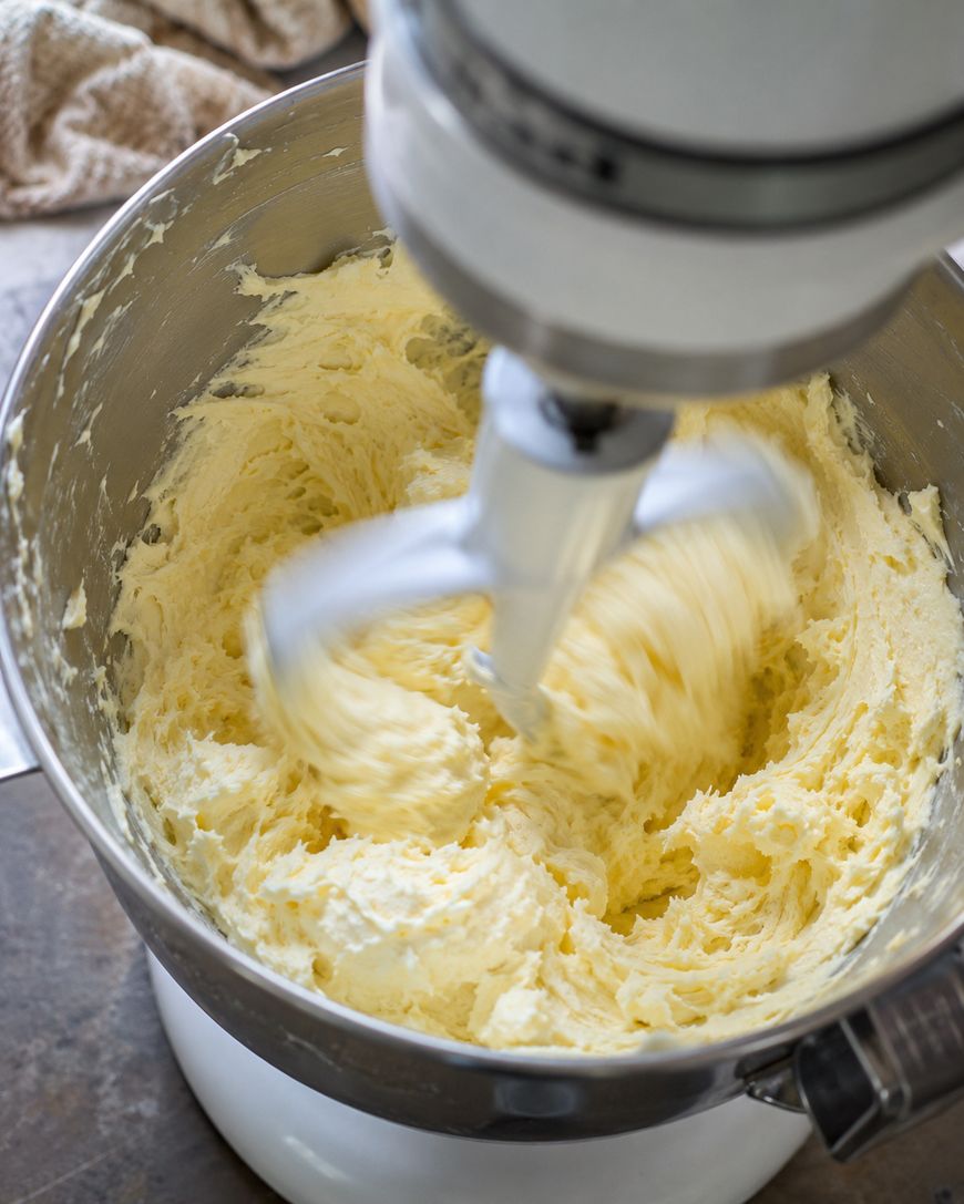 A stand mixer is creaming butter and sugar in a metal mixing bowl, making a fluffy, pale mixture. The paddle attachment is in motion, blending the ingredients. A beige towel is visible in the background.