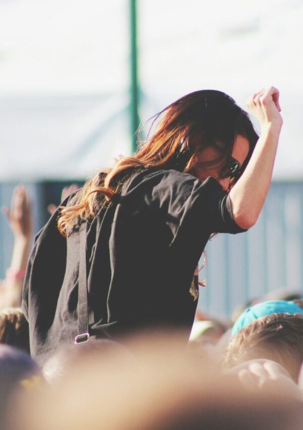 A woman wearing sunglasses and a black shirt dances and raises her arm while standing above a crowd at an outdoor event or concert in daylight.