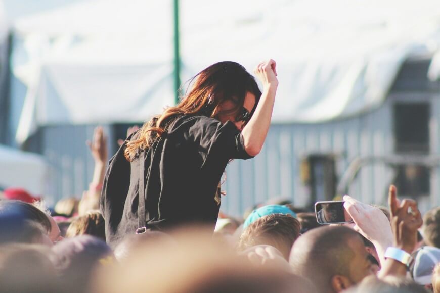 A woman wearing sunglasses sits on someone’s shoulders in a crowded outdoor event, raising her fist and enjoying the atmosphere. People around her are also watching and taking photos.